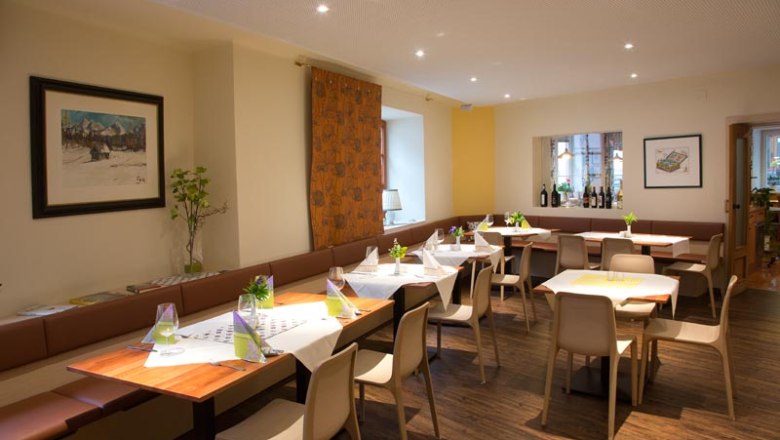 A modern dining room with laid tables, chairs and decorations. Pictures hang on the walls and there is a windowsill with bottles of wine.