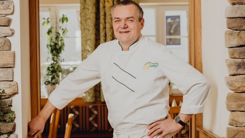 A man in a white chef's uniform stands smiling in a restaurant.