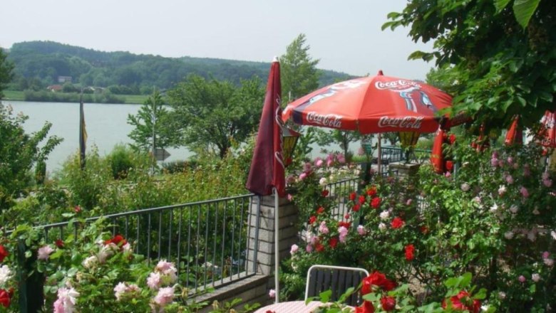 A blooming garden with red and pink roses, a table with chairs and a red parasol with the Coca-Cola logo, with a lake in the background.