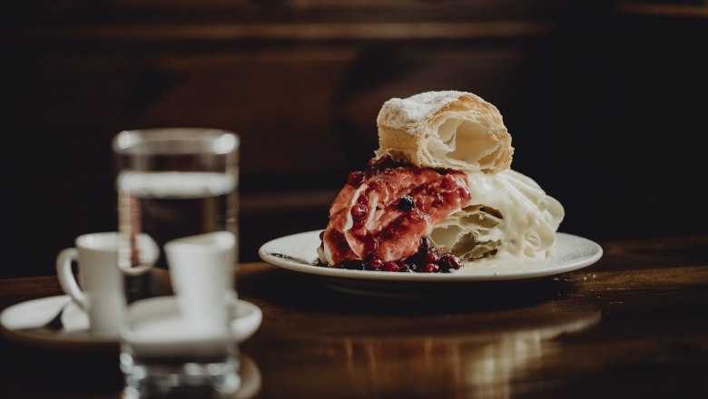 A plate with a monastery cream slice, garnished with red berries, stands on a wooden table. A glass of water and two espresso cups can be seen in the background.