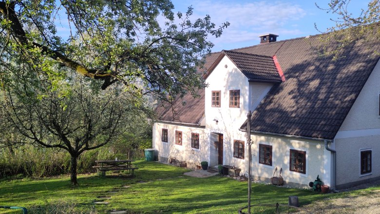 IMG_20260418_081143, © Andrea Stiftner A traditional country house with white walls and brown windows, surrounded by green trees and a well-tended garden with a picnic table.