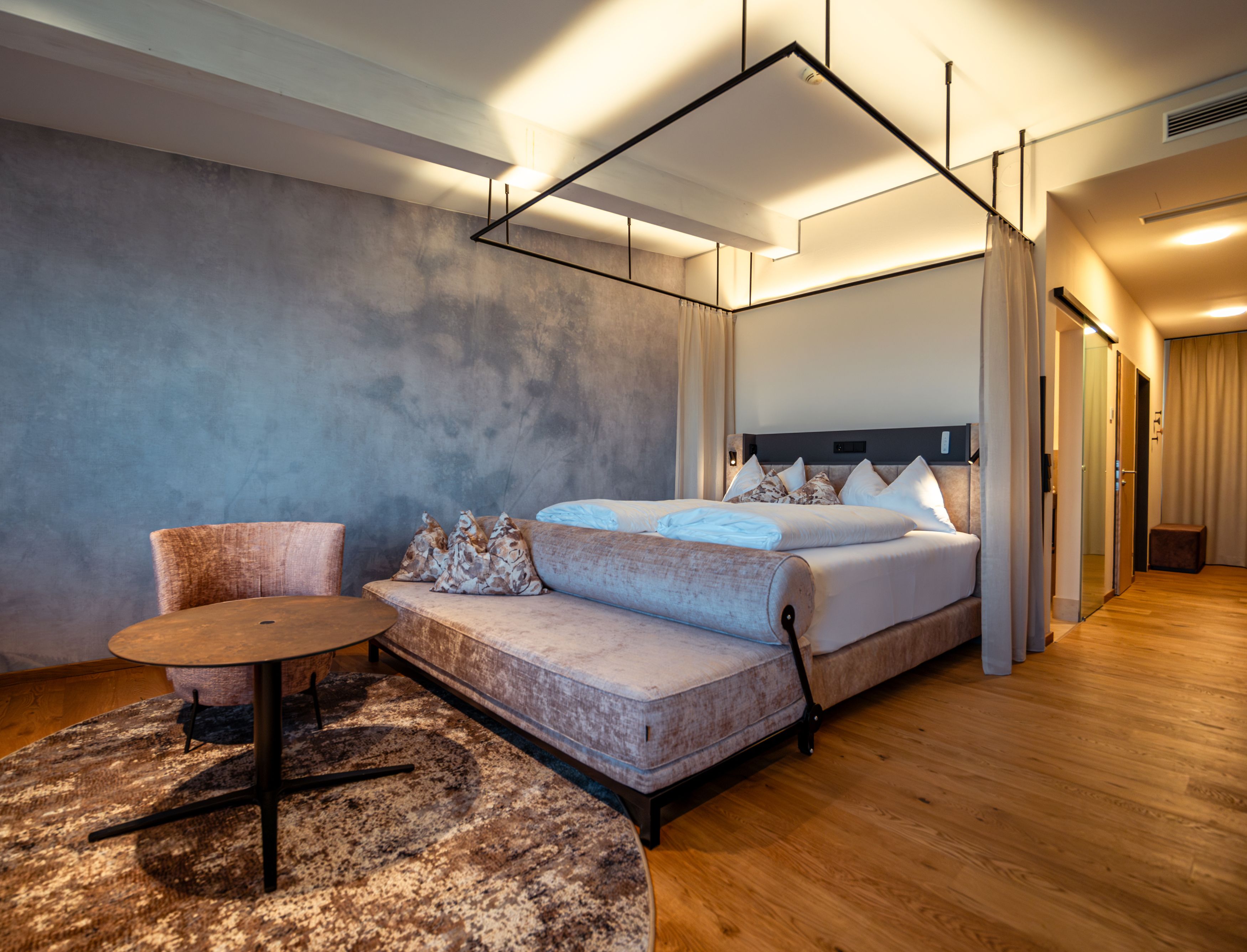 Modern hotel room with four-poster bed, armchair and table on wooden floor.