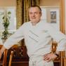A man in a white chef's uniform stands smiling in a restaurant.