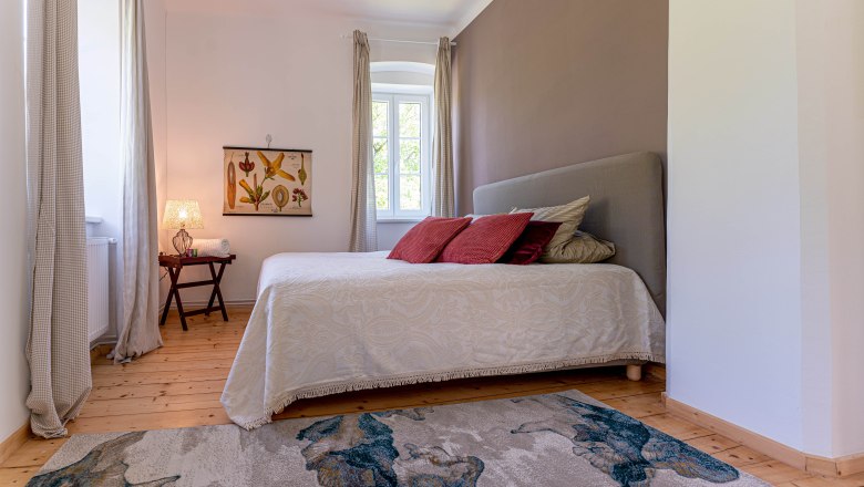 Bedroom with bed, red cushions, carpet and window with curtains.