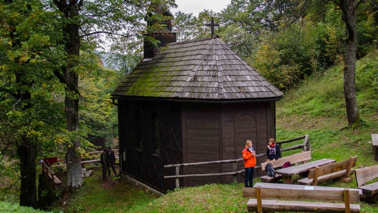 A small, wooden church in the forest with two people and wooden benches in the foreground.