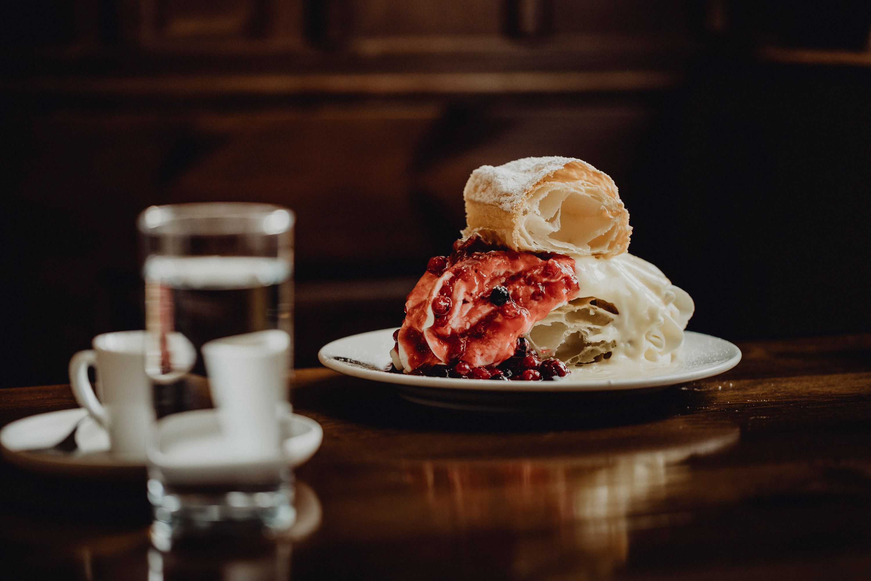 A plate with a monastery cream slice, garnished with red berries, stands on a wooden table. A glass of water and two espresso cups can be seen in the background.