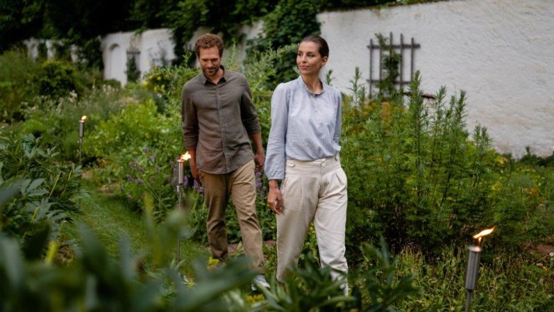 A man and a woman walk through a green garden surrounded by plants.