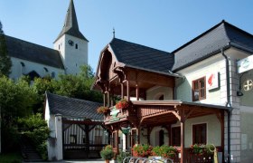 A traditional building with a wooden balcony and floral decorations, next to a church with a tower.