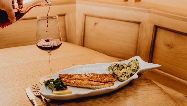 A plate with pike-perch fillet and potatoes in garlic butter, next to it a glass of red wine on a wooden table.