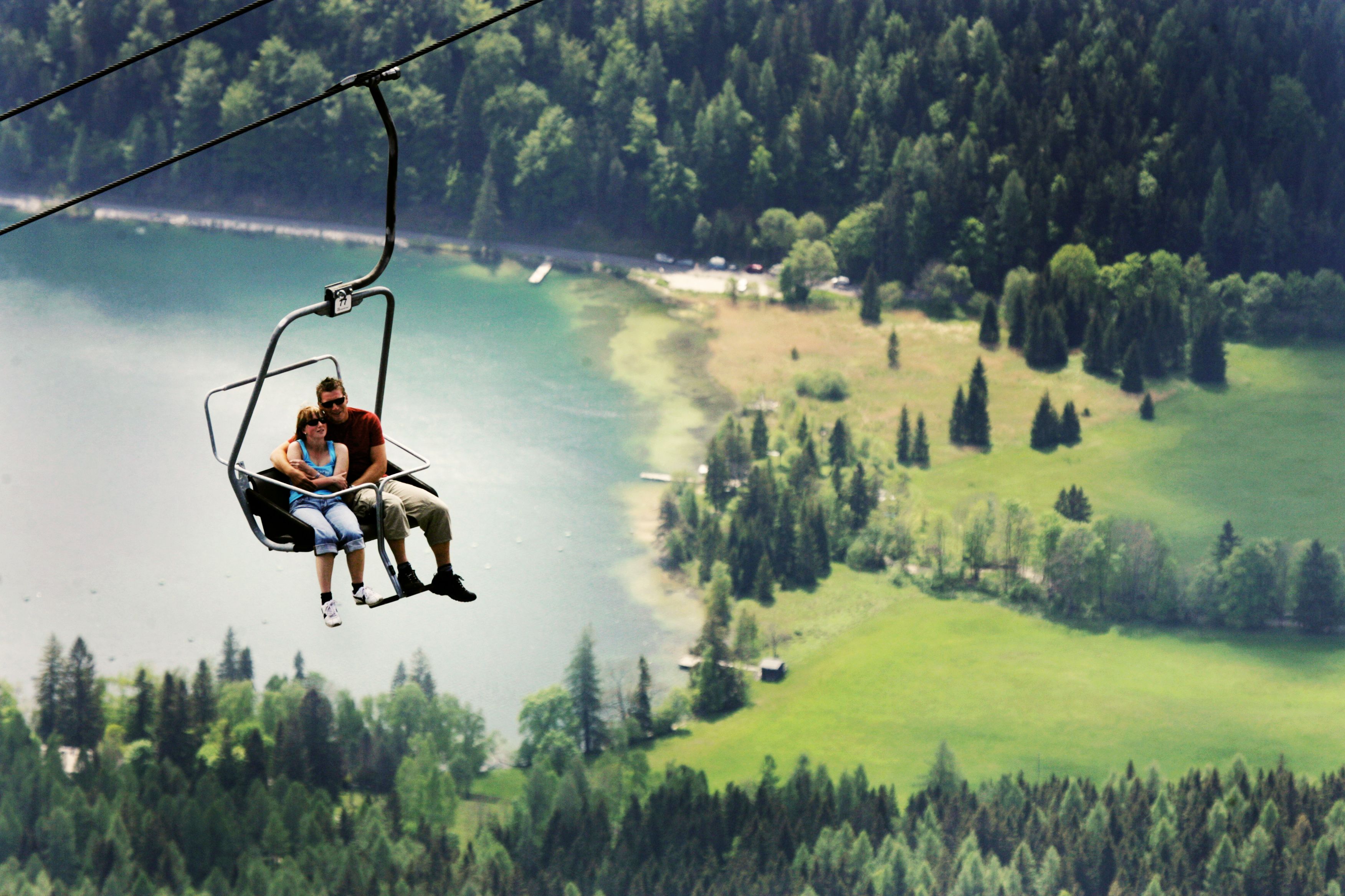 Two people sit in a chair lift above a green landscape with a lake in the background.