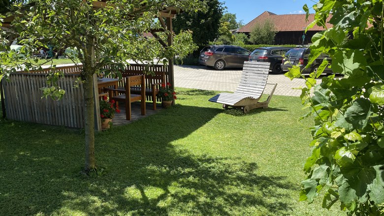Garden with pergola, wooden table and chairs, sun lounger on lawn, cars in the background.