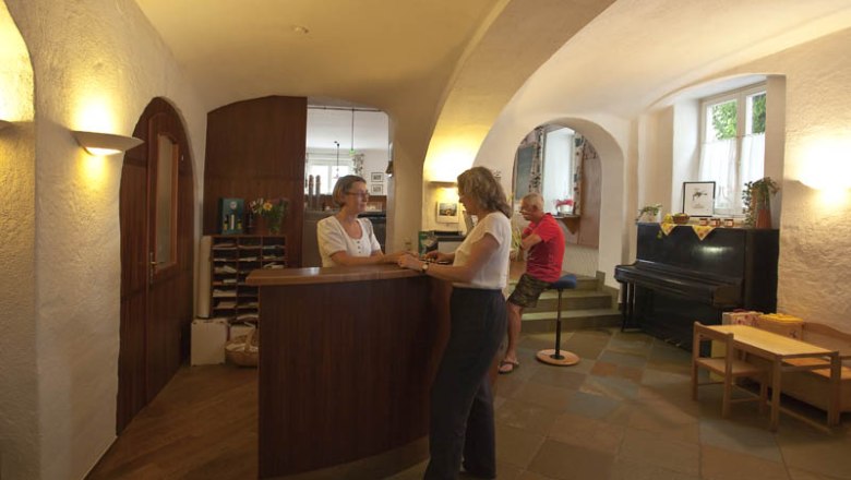 A hotel reception desk with two women in conversation and a man sitting on a stool.