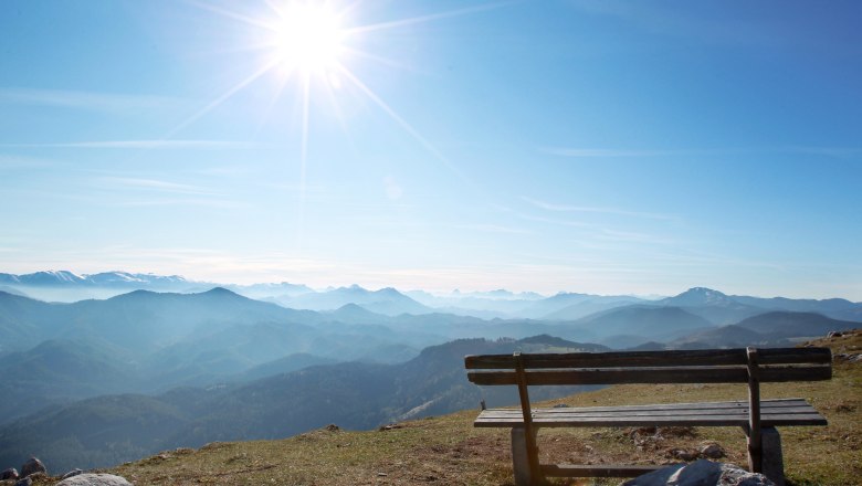 Wooden bench on a mountain with a view of a mountain landscape and the sun in the sky.