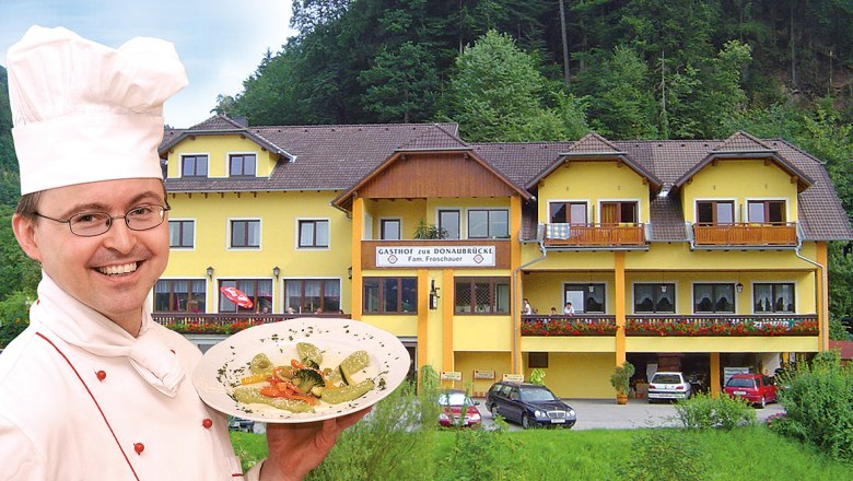 To the Danube bridge, © Helmut Froschauer A chef presents a dish in front of a yellow inn called 'Zur Donaubrücke'.