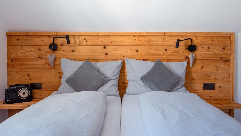 Comfortable double bed with wooden back panel, gray cushions and blankets. A retro radio on the left, a reading lamp and heart decoration on the right.
