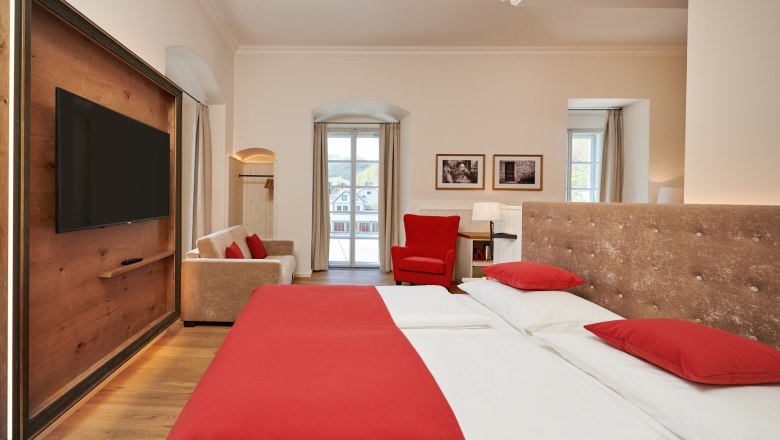 Hotel room with double bed, red bed linen, sofa, red armchair and large window.