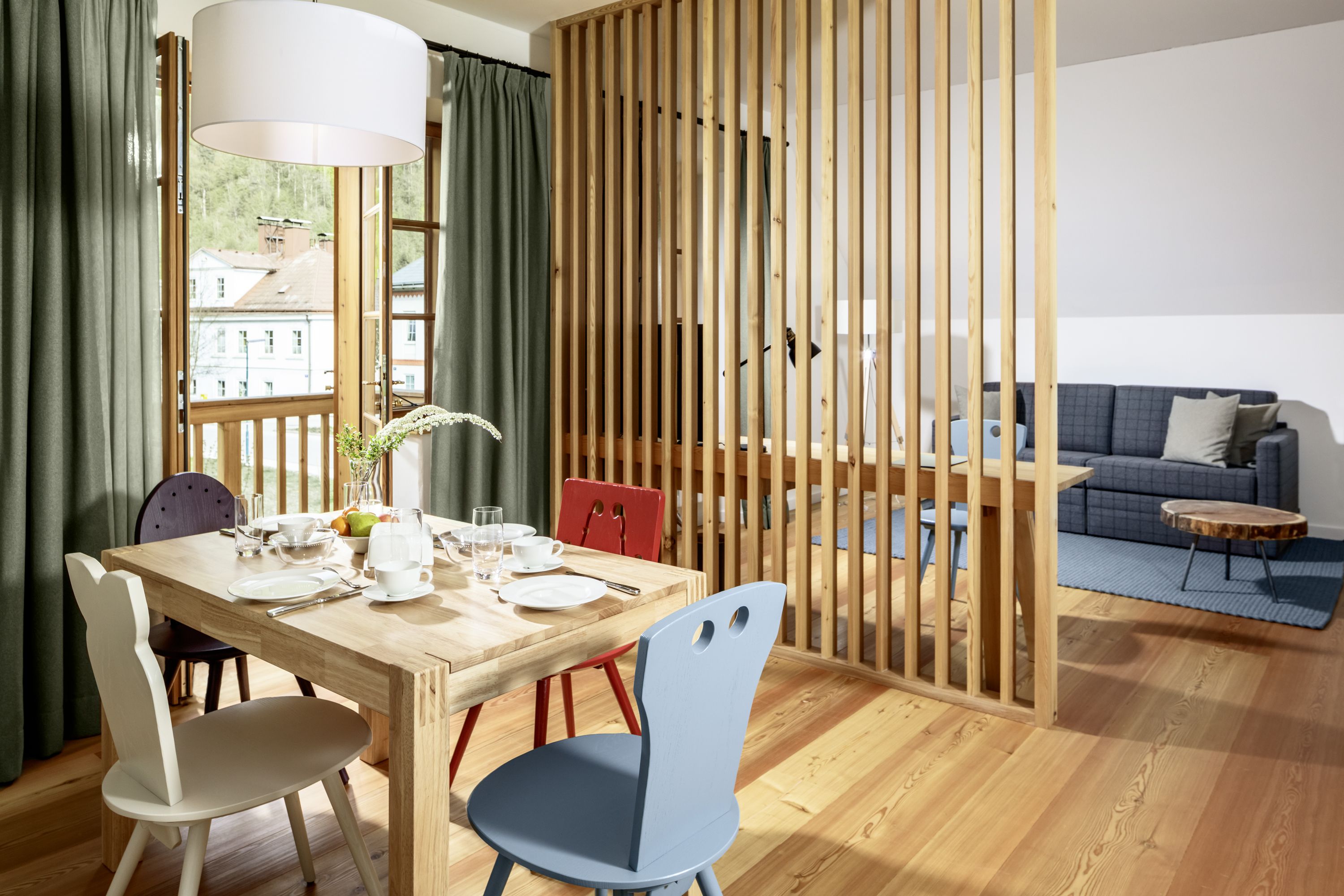 A bright hotel room with wooden floor, dining table and chairs, separated by a wooden slatted wall, with a view of a sofa and a coffee table.