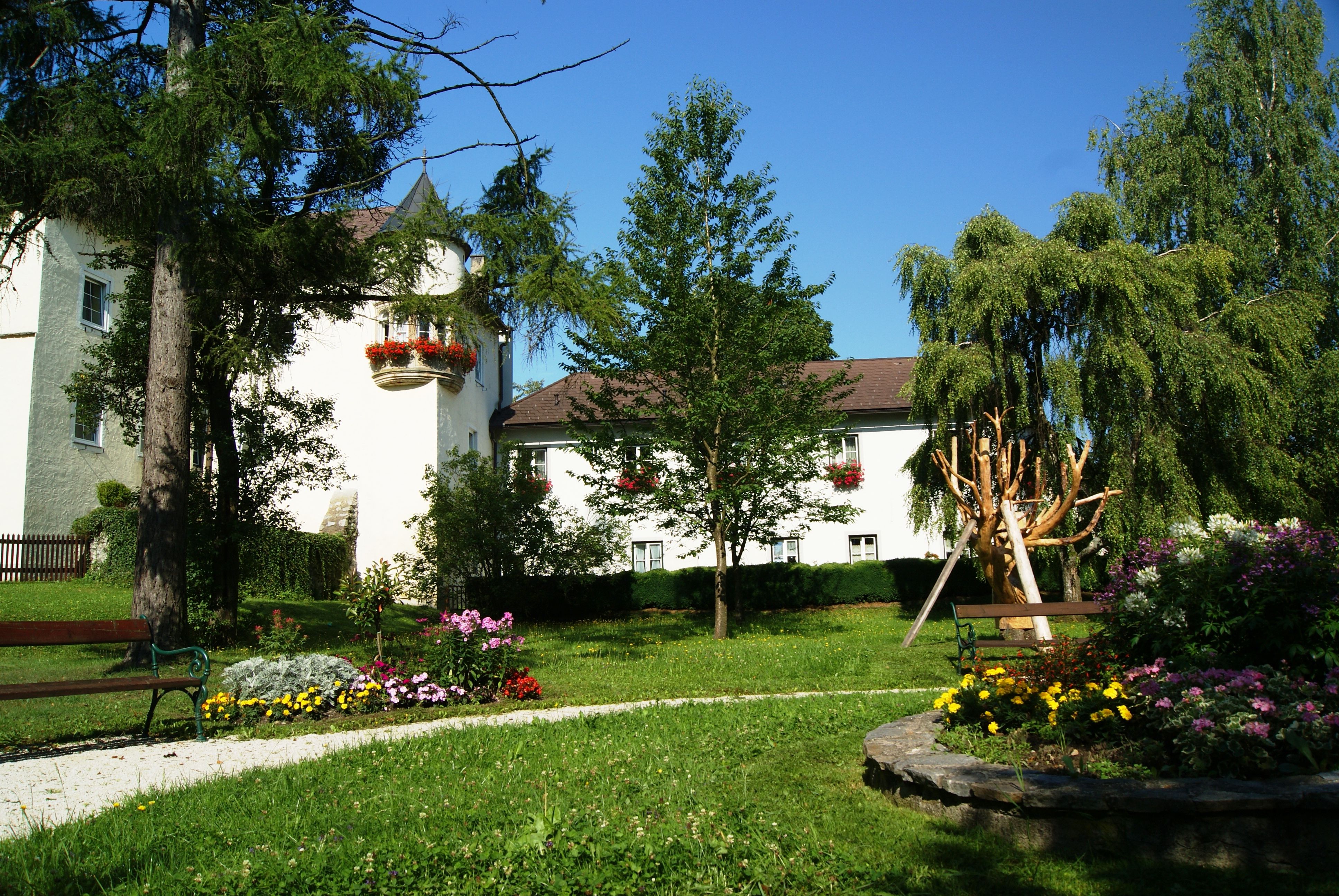 A castle with a white façade and red flowers, surrounded by a well-tended garden with trees and flowerbeds.
