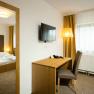 A hotel room with a double bed, a desk and chair, a TV on the wall and yellow curtains.