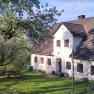A traditional country house with white walls and brown windows, surrounded by green trees and a well-tended garden with a picnic table.