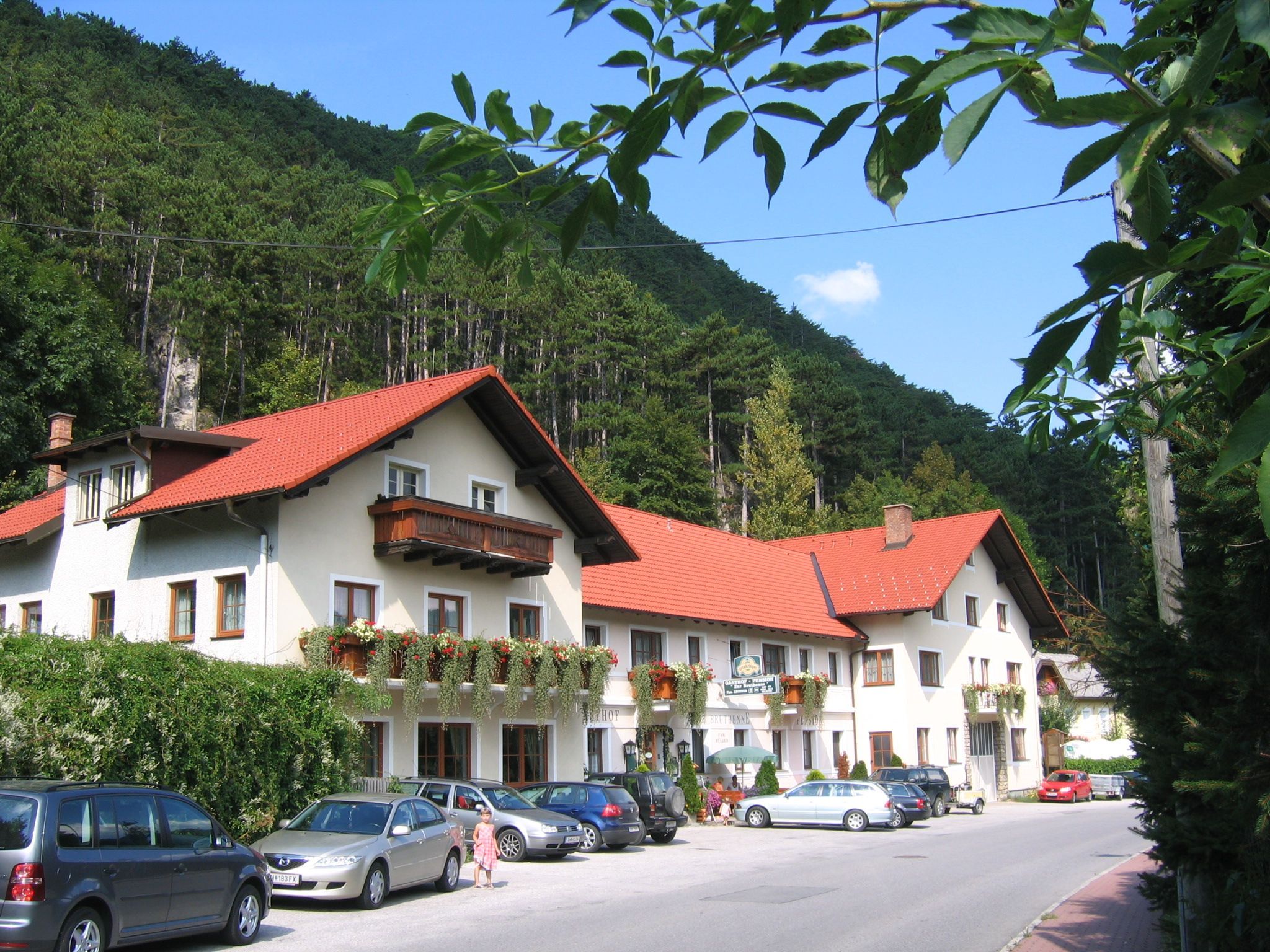 A traditional inn with red roofs and flower boxes, surrounded by woods and parked cars.