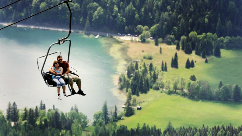 Two people sit in a chair lift above a green landscape with a lake in the background.