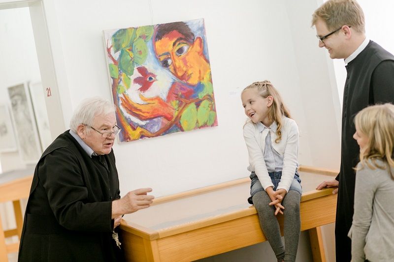 Two priests talk to two children in a gallery in front of a colorful painting.