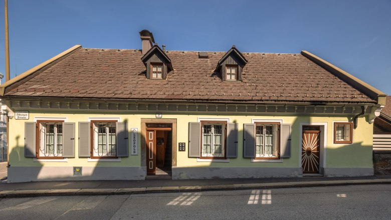 A traditional yellow house with a brown roof and green shutters on a street.