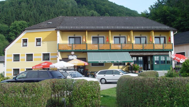 Yellow guesthouse with balcony and parasols in front of a wooded hill.