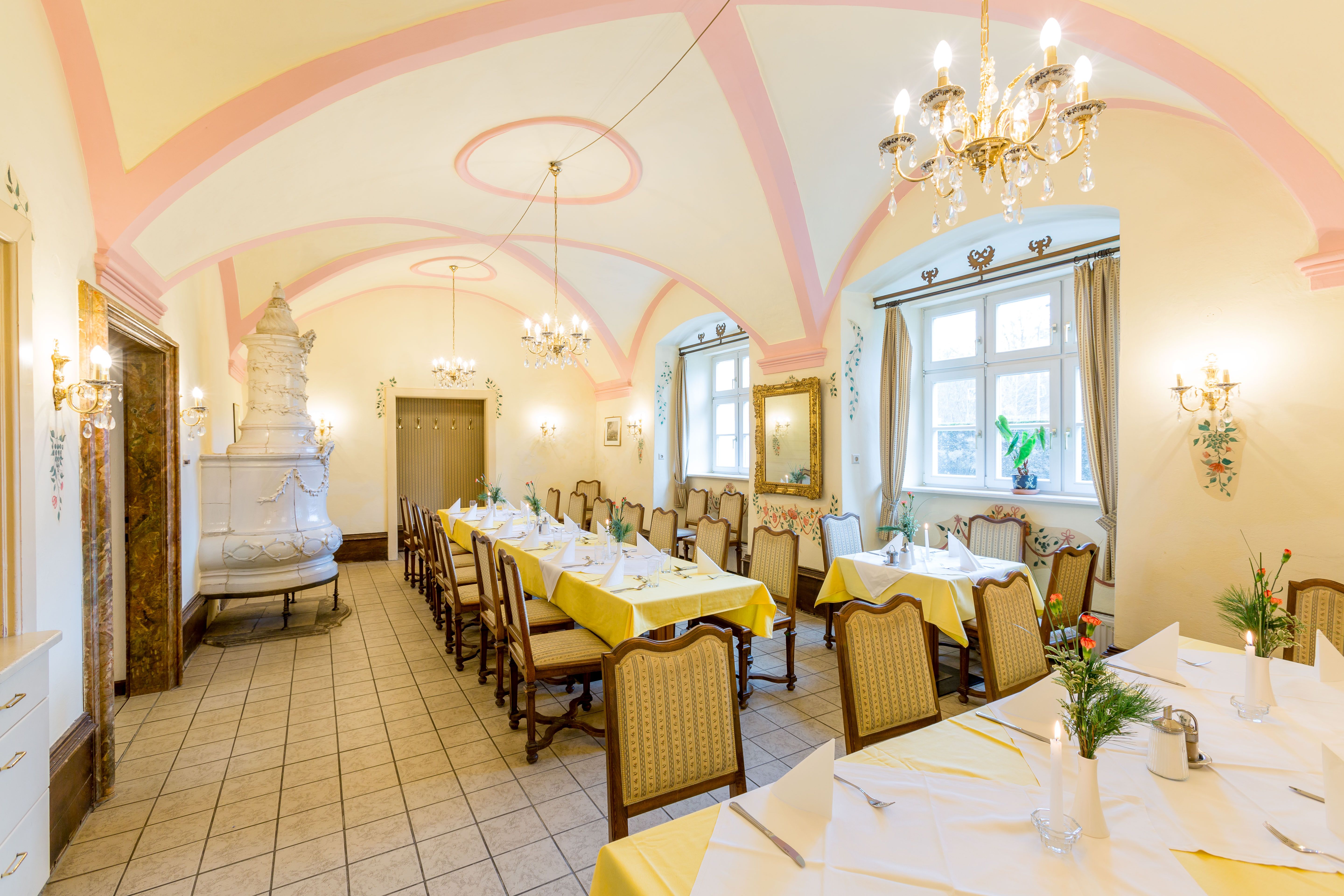Elegant dining room with laid tables, chandeliers and tiled stove.