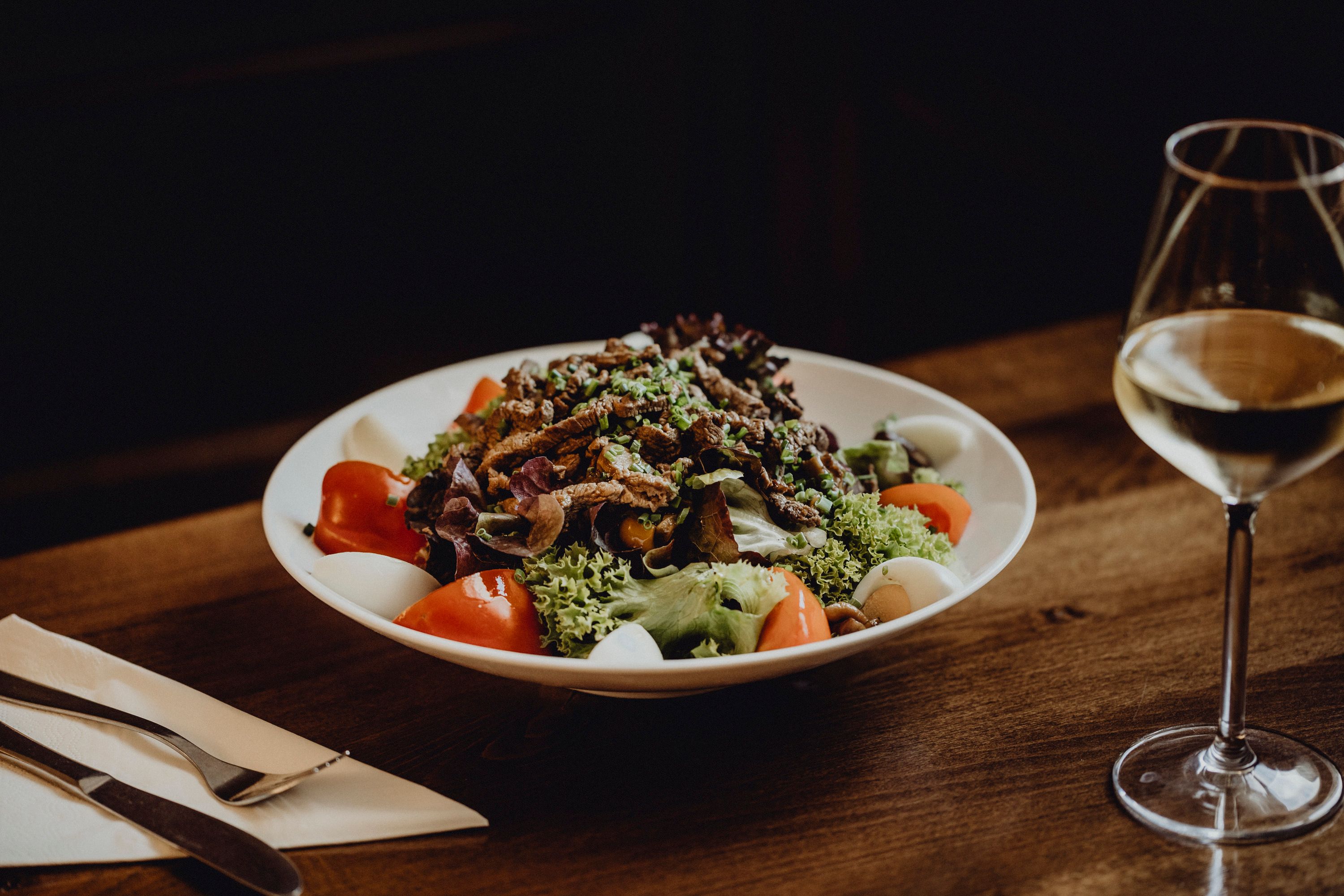 A plate of mixed salad, tomatoes, eggs and strips of meat on a wooden table, with a glass of white wine next to it.