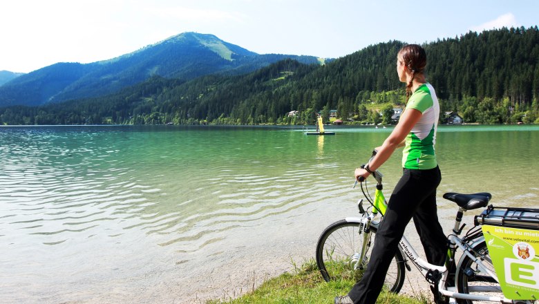 A woman with a bicycle stands on the shore of Lake Erlauf and looks out over the water and the surrounding mountains.