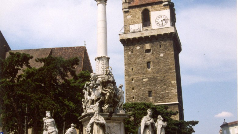 Historic defense tower with clock and baroque column in the foreground.