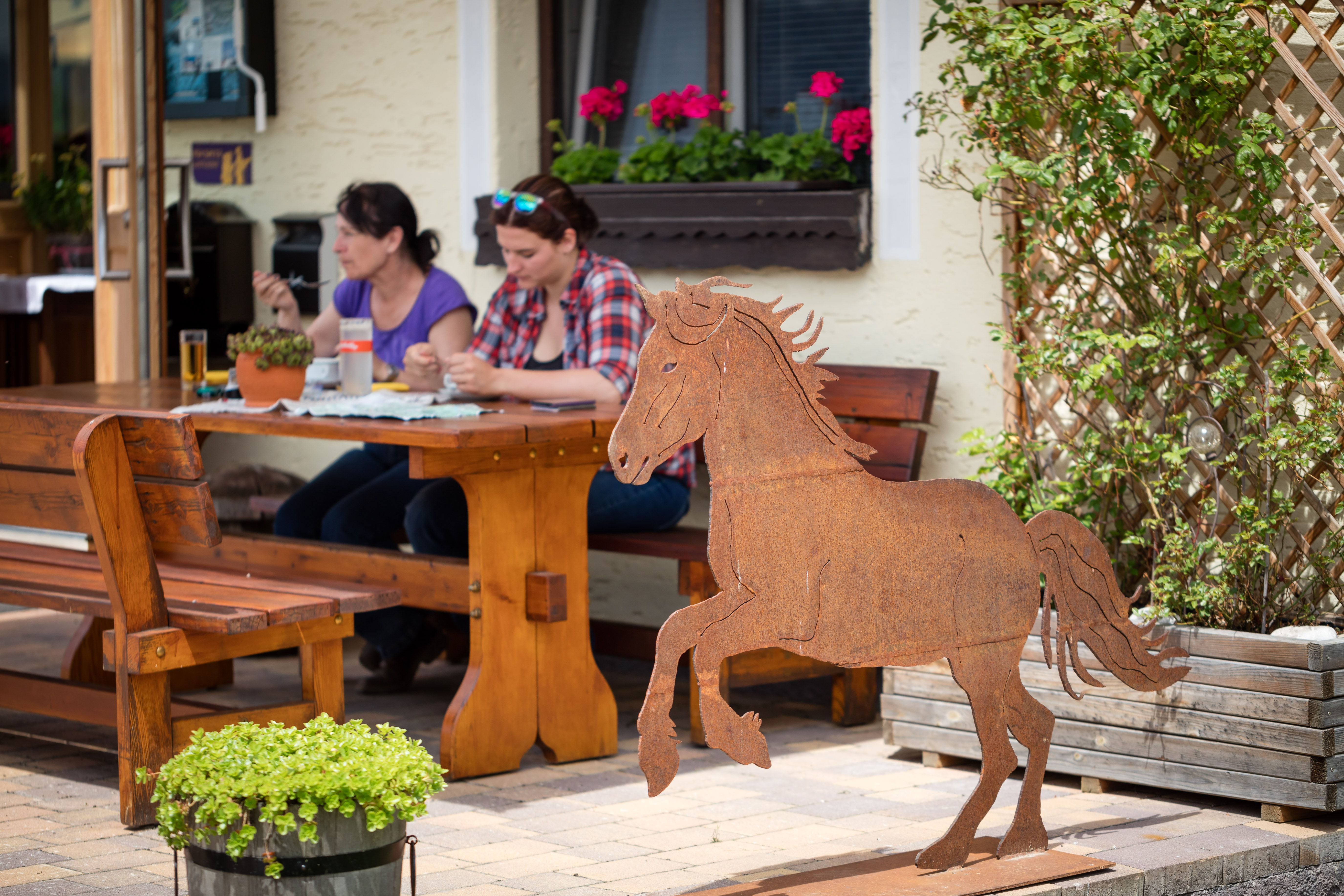 Two women sit at a wooden table outdoors and eat. In the foreground is a metal sculpture of a horse.