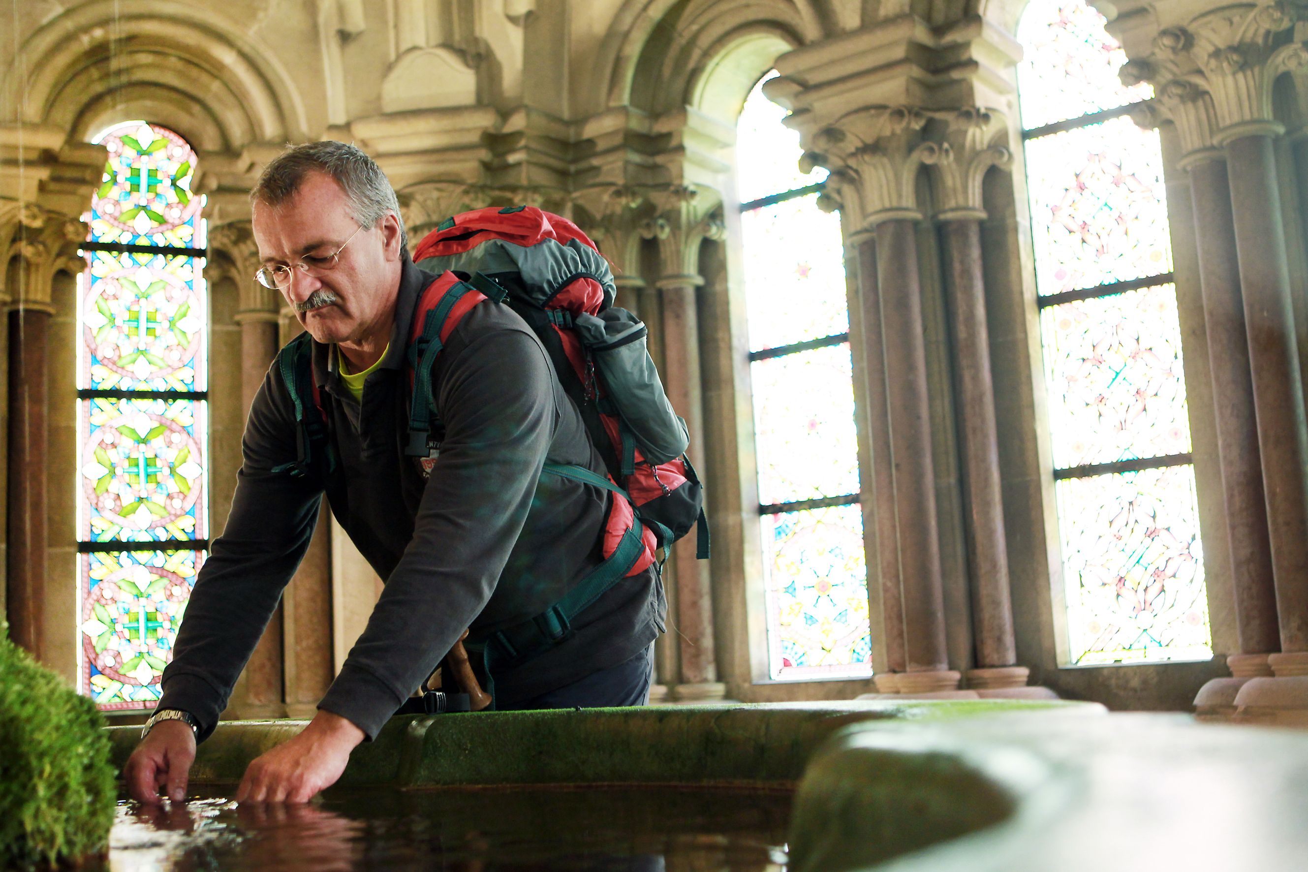 A man with a rucksack bends over a pool of water in a historic building with stained glass windows.