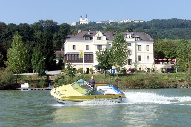 A yellow motorboat sails on a river in front of a large, light-colored building with hills in the background.