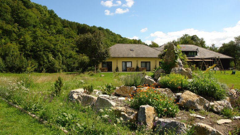 A yellow house with a garden and rocks in the foreground, surrounded by trees.