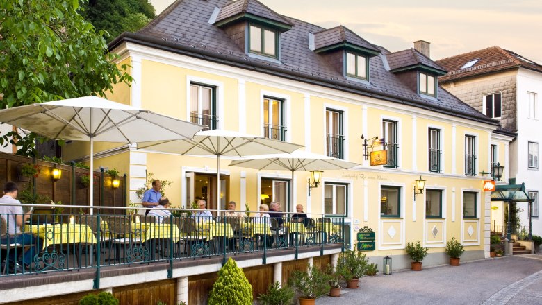 A yellow country inn with a terrace and parasols where people are sitting.
