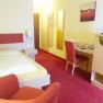 A hotel room with a single bed, red carpet, desk and chair. A picture hangs on the wall, next to it a television on a cupboard.