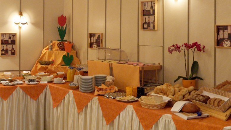 A rich breakfast buffet with bread, rolls, spreads and drinks on a table with an orange cloth.