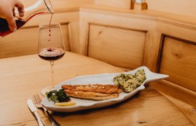 A plate with pike-perch fillet and potatoes in garlic butter, next to it a glass of red wine on a wooden table.