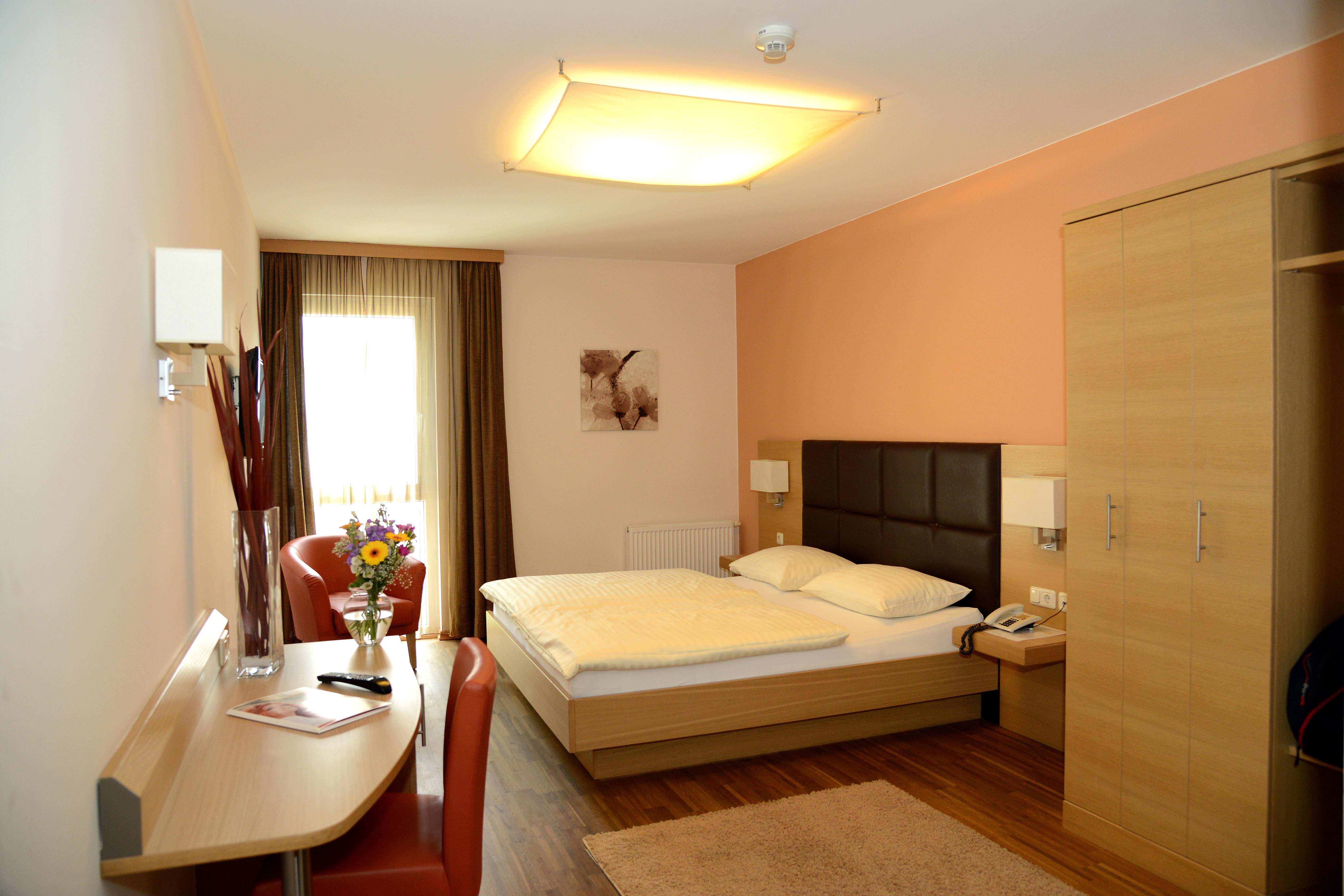 A modern hotel room with a double bed, desk, chair and bouquet of flowers on a table.