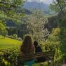 A woman is sitting on a bench with a dog, looking out at a green landscape with blossoming trees and mountains in the background.