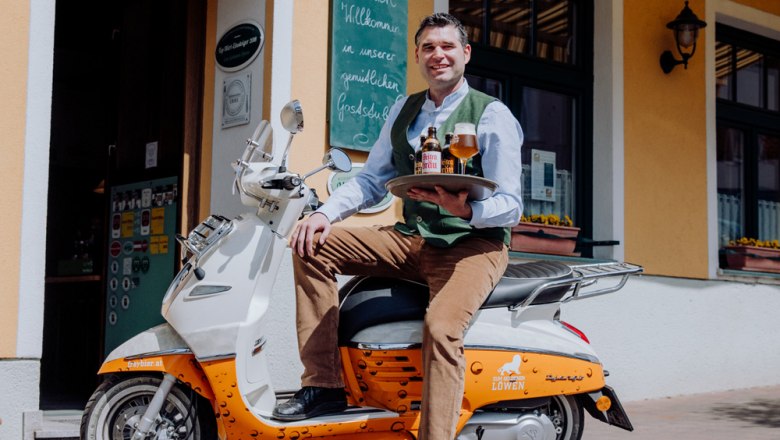 A man sits on a scooter in front of a building holding a tray with beer bottles and a glass.