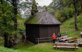 A small, wooden church in the forest with two people and wooden benches in the foreground.